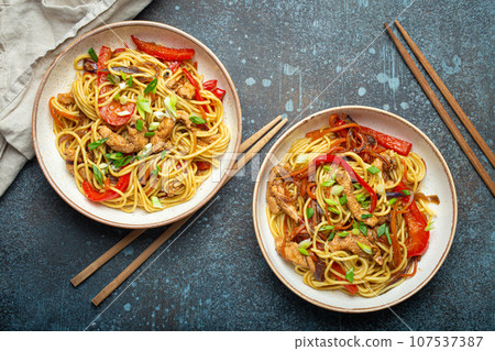 Two bowls with Chow Mein or Lo Mein, traditional Chinese stir fry noodles with meat and vegetables, served with chopsticks top view on rustic blue concrete background 107537387