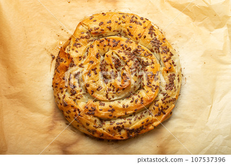 Freshly baked Burek made of filo dough with filling on baking paper background top view. Traditional savoury spiral pie of Balkans, Middle East and Central Asia Freshly baked Burek made of filo dough with filling on baking paper background top view. Traditional savoury spiral pie of Balkans, Middle East and Central Asia 107537396