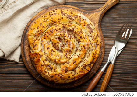 Burek made of phyllo dough with filling on cutting board, dark brown wooden rustic background top view. Traditional savoury spiral pie of Balkans, Middle East and Central Asia Burek made of phyllo dough with filling on cutting board, dark brown wooden rustic background top view. Traditional savoury spiral pie of Balkans, Middle East and Central Asia 107537399