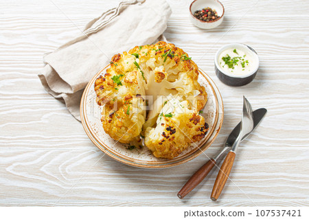 Whole roasted head of cauliflower with olive oil on plate on white wooden rustic table top view with cutlery. Vegetarian dish. Whole roasted head of cauliflower with olive oil on plate on white wooden rustic table top view with cutlery. Vegetarian dish. 107537421