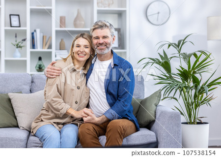 Portrait of mature gray-haired couple, happy family man and woman smiling and looking at camera, sitting on sofa in living room at home. 107538154