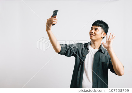 Smiling Asian man captures the moment with a selfie on his smartphone in a studio shot on white background. Self-expression and fun photography, hello to the world Smiling Asian man captures the moment with a selfie on his smartphone in a studio shot on white background. Self-expression and fun photography, hello to the world 107538799