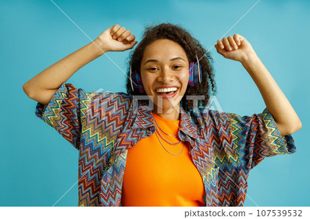 Happy woman in headphones dancing with raised hands while standing on blue studio background Happy woman in headphones dancing with raised hands while standing on blue studio background 107539532
