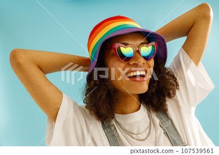 Smiling woman in rainbow cap and heart-shaped glasses looks away over blue studio background 107539561