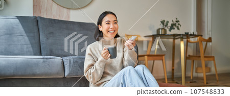 Portrait of beautiful asian girl sitting at her home and watching tv, holding remote, smiling and laughing, feeling comfort and warmth at her apartment Portrait of beautiful asian girl sitting at her home and watching tv, holding remote, smiling and laughing, feeling comfort and warmth at her apartment 107548833