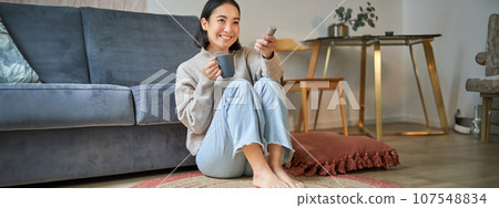 Portrait of beautiful asian girl sitting at her home and watching tv, holding remote, smiling and laughing, feeling comfort and warmth at her apartment Portrait of beautiful asian girl sitting at her home and watching tv, holding remote, smiling and laughing, feeling comfort and warmth at her apartment 107548834