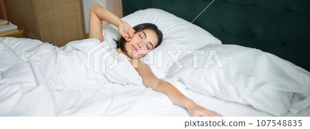 Romantic asian girl waking up in cozy bedroom, lying in bed with white sheets, stretching her hand towards empty pillow Romantic asian girl waking up in cozy bedroom, lying in bed with white sheets, stretching her hand towards empty pillow 107548835