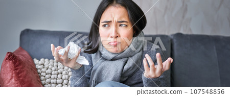 Cost of living and heating problems. Close up of angry girl sneezing, staying at home and freezing, wearing warm clothes and scarf 107548856