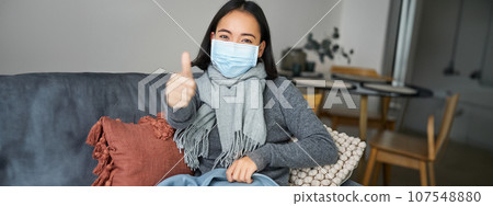 Covid-19 and health concept. Young asian woman in medical face mask, feels sick and unwell, catching flu, protecting others from influenza, sitting in living room covered with blanket Covid-19 and health concept. Young asian woman in medical face mask, feels sick and unwell, catching flu, protecting others from influenza, sitting in living room covered with blanket 107548880