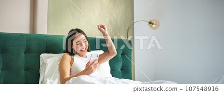 Enthusiastic asian girl reading mobile phone news, raising hand and triumphing in her bed, cheering while lying on pillow in bedroom, positive news 107548916