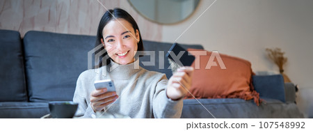 Smiling cute asian woman using credit card and smartphone, paying bills online, holding mobile phone, looking at camera 107548992