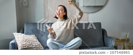 Portrait of carefree asian woman singing and listening music from smartphone app, using wireless headphones, smiling pleased, sitting on sofa at home 107548993