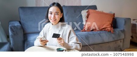 Smiling cute asian woman using credit card and smartphone, paying bills online, holding mobile phone, looking at camera 107548998