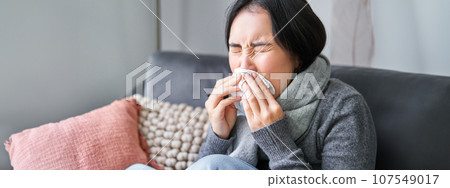 Close up of young korean woman staying at home with cold, sneezing in napking, has runny nose, concept of illness, health and influenza Close up of young korean woman staying at home with cold, sneezing in napking, has runny nose, concept of illness, health and influenza 107549017