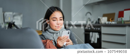Portrait of ill young korean woman sitting on sofa, texting message, using mobile phone to contact her doctor while being sick 107549038