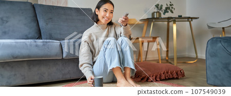Portrait of beautiful asian girl sitting at her home and watching tv, holding remote, smiling and laughing, feeling comfort and warmth at her apartment Portrait of beautiful asian girl sitting at her home and watching tv, holding remote, smiling and laughing, feeling comfort and warmth at her apartment 107549039