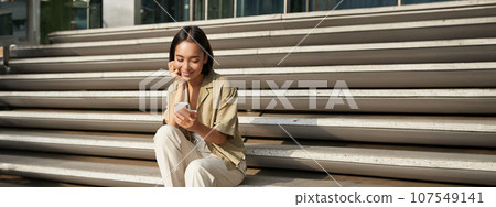 Vertical shot of asian woman, student sits on stairs in city, looking at mobile phone screen and smiling, using smartphone app Vertical shot of asian woman, student sits on stairs in city, looking at mobile phone screen and smiling, using smartphone app 107549141