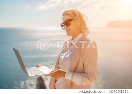 Freelance women sea. She is working on the computer. Good looking middle aged woman typing on a laptop keyboard outdoors with a beautiful sea view. The concept of remote work. 107553981
