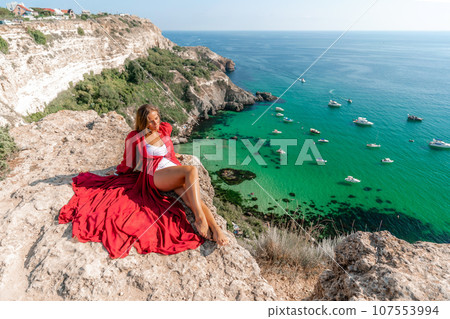 Woman red dress sea. Happy woman in a red dress and white bikini sitting on a rocky outcrop, gazing out at the sea with boats and yachts in the background. 107553994
