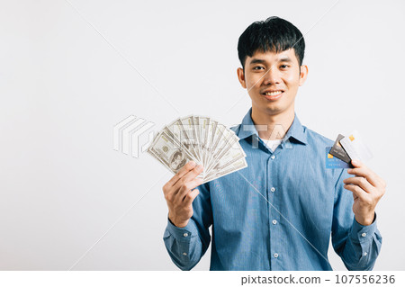 Smiling man holds dollars and a credit card, symbolizing financial success and payment options. Studio shot isolated on white, showcasing his confidence in financial matters. 107556236