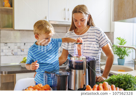 Little child boy with mother makes fresh carrot juice with juicer in kitchen 107562328