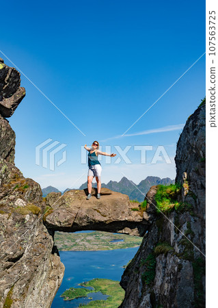 Brave traveler woman standing on hanging stone between rocks. Djevelporten in Norway Lofoten islands Brave traveler woman standing on hanging stone between rocks. Djevelporten in Norway Lofoten islands 107563725
