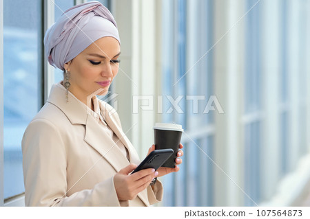 Muslim woman drinks beverage scrolling social media by office Muslim woman drinks beverage scrolling social media by office 107564873