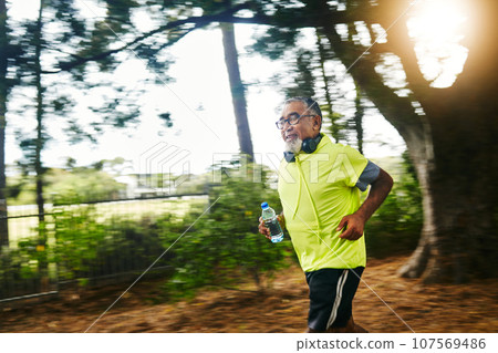 Senior man, running and listening to music on road in nature, countryside or forest with water bottle in exercise. Fitness, training and workout or speed run in woods for cardio, health and wellness Senior man, running and listening to music on road in nature, countryside or forest with water bottle in exercise. Fitness, training and workout or speed run in woods for cardio, health and wellness 107569486