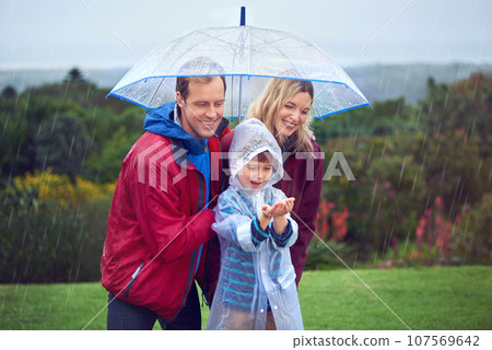 Rain, umbrella and a happy family outdoor in nature for fun, happiness and quality time. Man, woman and excited child together with hands to catch water drops while learning, playing or development 107569642