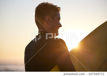 Fitness, sunset or man surfer with surfboard for exercise, workout or training wellness at the beach, ocean or sea with smile. Happy, freedom or lens flare male athlete for water sports motivation 107569731