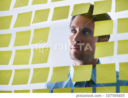 Businessman, face and thinking in planning, sticky note or brainstorming tasks on glass board at office. Thoughtful man contemplating business decision, strategy or schedule in wonder at workplace 107569885