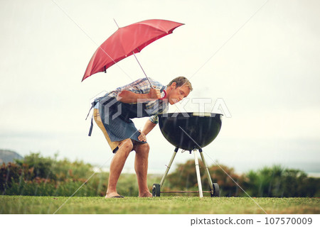 Umbrella, rain and a man outdoor to barbecue food for cooking or insurance in the winter season. Storm, weather and grill with a male person getting wet while trying to bbq on a grass lawn or garden 107570009