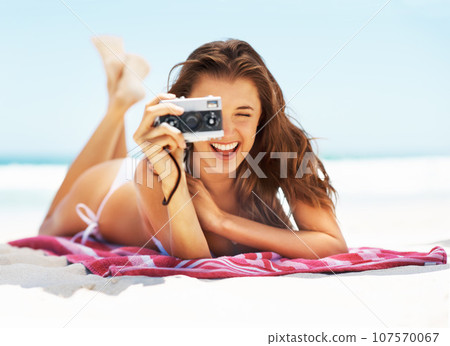 Portrait, camera and travel with a woman on the beach during summer for a holiday or luxury vacation. Ocean photo, bikini and smile with a happy young tourist on the sand to relax by the coastal sea 107570067