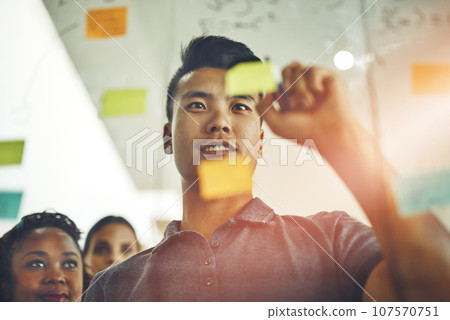 A marketing professional brainstorming ideas with colleagues, writing on transparent board with sticky notes during meeting. Young, Asian entrepreneur discussing a work project schedule or timeline. A marketing professional brainstorming ideas with colleagues, writing on transparent board with sticky notes during meeting. Young, Asian entrepreneur discussing a work project schedule or timeline. 107570751