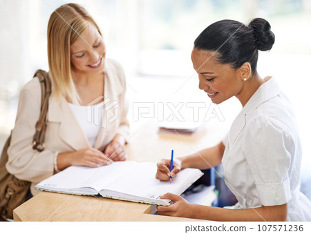 Woman signing in with a receptionist at a desk for an appointment, meeting or reservation. Happy, conversation and secretary writing in a book for checking in at counter with female person in office. Woman signing in with a receptionist at a desk for an appointment, meeting or reservation. Happy, conversation and secretary writing in a book for checking in at counter with female person in office. 107571236