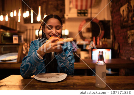 Pizza, food and restaurant where young woman is eating food and enjoying drinks. Hungry female having delicious meal at bar at night. Happy and satisfied foodie having tasty supper at a diner Pizza, food and restaurant where young woman is eating food and enjoying drinks. Hungry female having delicious meal at bar at night. Happy and satisfied foodie having tasty supper at a diner 107571828