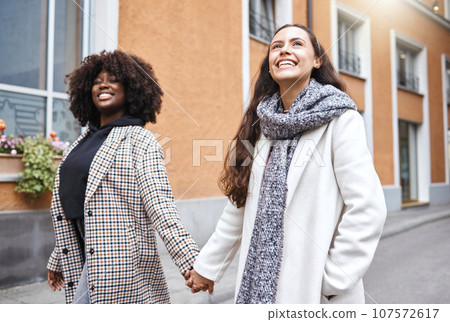 Woman, friends and holding hands walking in the city for friendship, travel or fun journey in the outdoors. Happy women taking walk or stroll touching hand with smile for sightseeing in an urban town 107572617