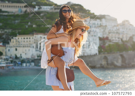 Vacation piggyback, happy and women by the water for freedom, adventure and walking in Spain. Smile, travel and young friends being carefree together on a holiday in a small town by the ocean 107572793