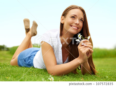 Thinking, smile and woman with daisy on grass, lying down on field and enjoying spring on vacation outdoor. Happy, flower plant and beauty of female person relaxing, smiling and having fun in nature. 107572835