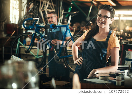 Portrait, working and smile of woman in bicycle shop, repair store or cycling workshop. Face, bike mechanic and confident person, professional or technician standing with glasses in small business. Portrait, working and smile of woman in bicycle shop, repair store or cycling workshop. Face, bike mechanic and confident person, professional or technician standing with glasses in small business. 107572952