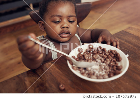 Black child, cereal and eating baby in a home kitchen with food and bowl at breakfast. African girl, nutrition and youth in a house with hungry kid relax with chocolate fiber snack in the morning Black child, cereal and eating baby in a home kitchen with food and bowl at breakfast. African girl, nutrition and youth in a house with hungry kid relax with chocolate fiber snack in the morning 107572972