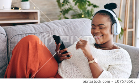 Black woman on couch, headphones for music and smartphone, relax at home and audio streaming with smile. Happiness, mobile app and using phone for radio or podcast, scroll with technology in lounge 107576503