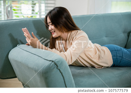 Smiling attractive woman relaxed reading text message on smartphone, Asian young female using smart mobile phone lying on sofa in living room room at home, gadget online application making internet 107577579