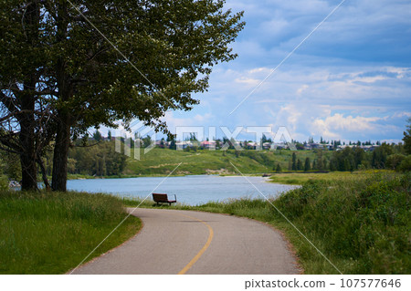 A path in a beautiful summer city park by the river. A bench on the shore of a calm lake in the forest. 107577646