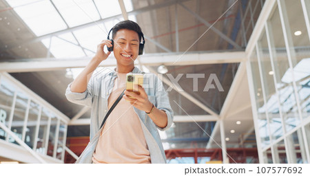 Walking, phone or Asian man in office with headphones on social media, internet or network in a building. Podcast, travel or happy intern on mobile app to chat or scroll for radio music playlist 107577692