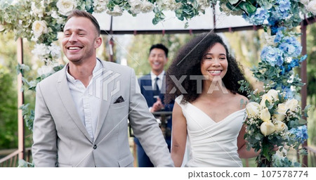 Couple, wedding and walk with flower confetti for event, celebration and outdoor in nature. Girl, happy interracial marriage and floral bouquet in park, holding hands or fist in air, pride or party 107578774