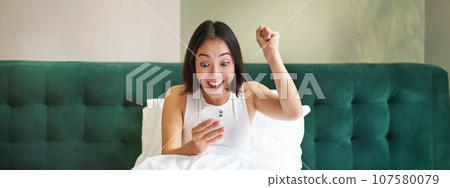 Happy asian girl reading on mobile phone and celebrating. Korean woman in bed, looking at smartphone and triumphing Happy asian girl reading on mobile phone and celebrating. Korean woman in bed, looking at smartphone and triumphing 107580079