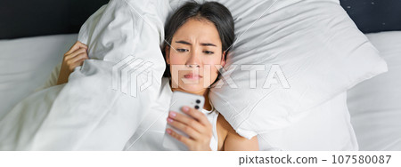 Close up portrait of asian girl lying in bed, looking at smartphone concerned, waking up late and staring at her alarm clock on mobile phone 107580087