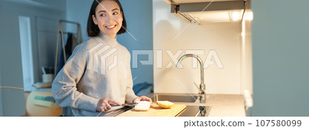 Cute asian woman making herself toast, cut loaf of bread, preparing sandwitch on kitchen Cute asian woman making herself toast, cut loaf of bread, preparing sandwitch on kitchen 107580099