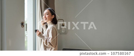 Beautiful korean woman staying at home, looking outside window, drinking coffee espresso and smiling, feeling comfort and warmth Beautiful korean woman staying at home, looking outside window, drinking coffee espresso and smiling, feeling comfort and warmth 107580167